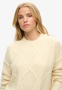 Sweter w kolorze kremowym o teksturowanym splocie, z diamentowym wzorem oraz ściągaczami przy dekolcie i mankietach. Miękka, gruba tkanina.