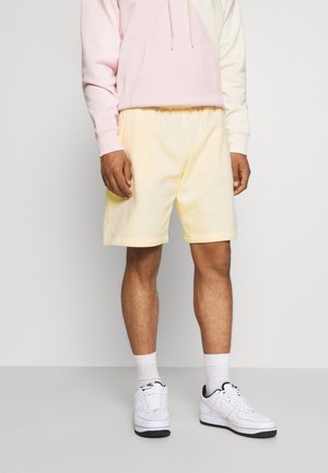 Persoon die lichtgele shorts, witte sportsokken, witte sneakers en een lichtroze-wit lange mouwen hoodie draagt, staand tegen een eenvoudige achtergrond.