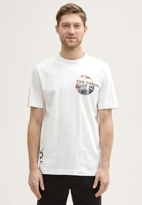 Hvid bomulds T-shirt med korte ærmer, med et cirkulært grafisk print i sort og beige tekst. Sorte detaljer på kanten.