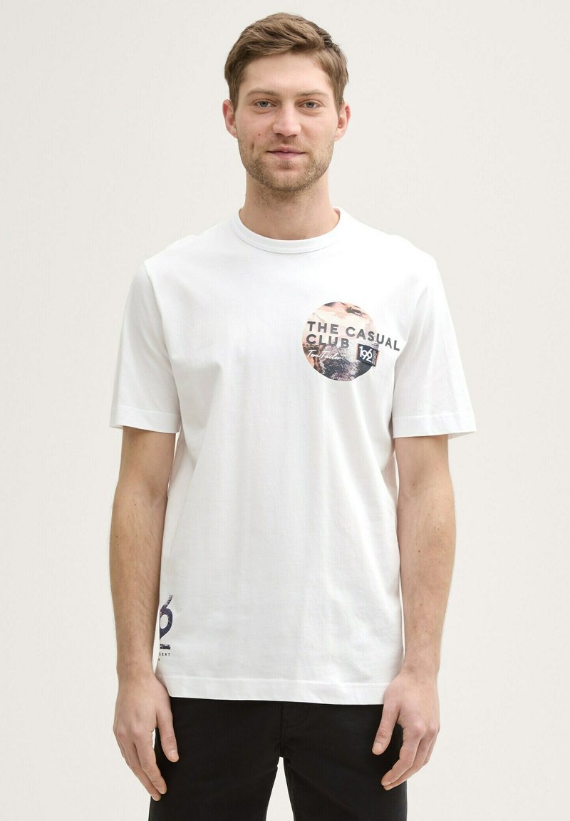 Hvid bomulds T-shirt med korte ærmer, med et cirkulært grafisk print i sort og beige tekst. Sorte detaljer på kanten.