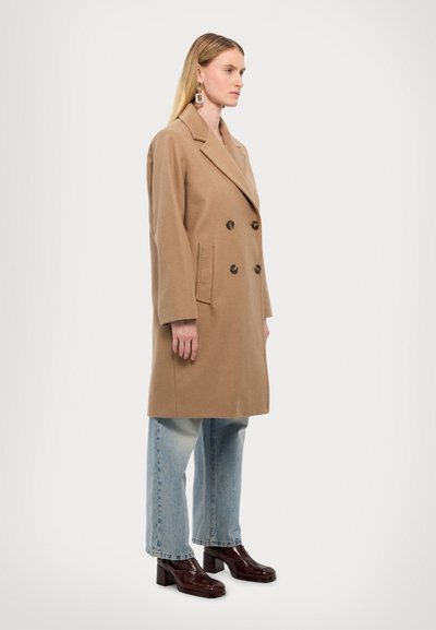 Manteau beige à double boutonnage avec de grands boutons, deux poches avant et une coupe décontractée. Associé à un jean large bleu clair et des chaussures à plateforme foncées.
