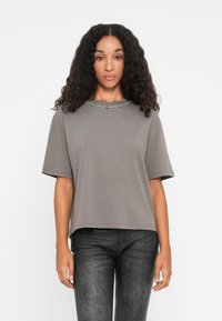 Grå t-skjorte med kort erm og rund halskant pyntet med metalliske detaljer, sammen med mørke strukturerte jeans.