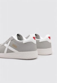 Zapatillas gris con un acabado suave, detalles en blanco, suelas texturizadas y un detalle en rojo en el talón. Incluyen un logo en el lateral y cordones planos.