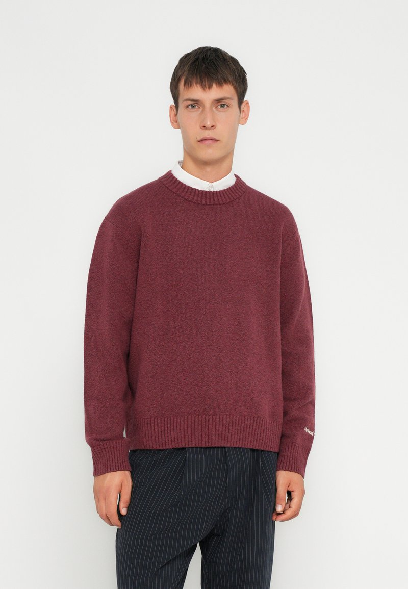 Suéter de punto burdeos con un cuello redondo acanalado y puños, que presenta una textura suave y un corte holgado, combinado con pantalones rayados.