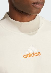 Šviesiai smėlinė sportinė palaidinė su aukštu raukšlėtu apykaklės kraštu. Pasižymi išsiuvinėtu baltu ir oranžiniu adidas logotipu ant krūtinės. Lygus paviršius.