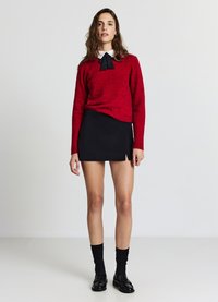Roter Strickpullover mit rundem Ausschnitt und schwarzer Schleife über einem weißen Hemd mit Kragen, kombiniert mit einem schwarzen Minirock und schwarzen Socken.
