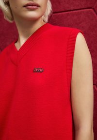 Red sleeveless knit vest with a V-neck, featuring ribbed edges and a small blue and red logo on the front. Textured fabric detail visible.
