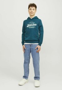 Felpa in cotone color turchese con stampa bianca, abbinata a jeans in denim blu chiaro e sneakers bianche. Il modello è in piedi contro uno sfondo neutro.