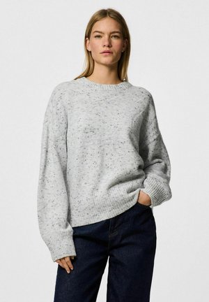 Maglione grigio a maglia con una texture maculata, colletto rotondo e spalle scese, abbinato a jeans scuri. Vestibilità rilassata e polsini a coste.