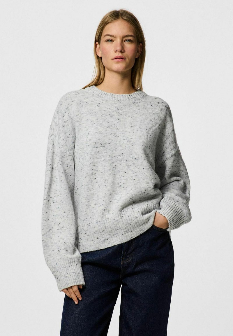 Grå stickad tröja med en fläckig textur, rund halsringning och sänkte axlar, kombinerad med mörka jeans. Avslappnad passform och ribbade ärmslut.