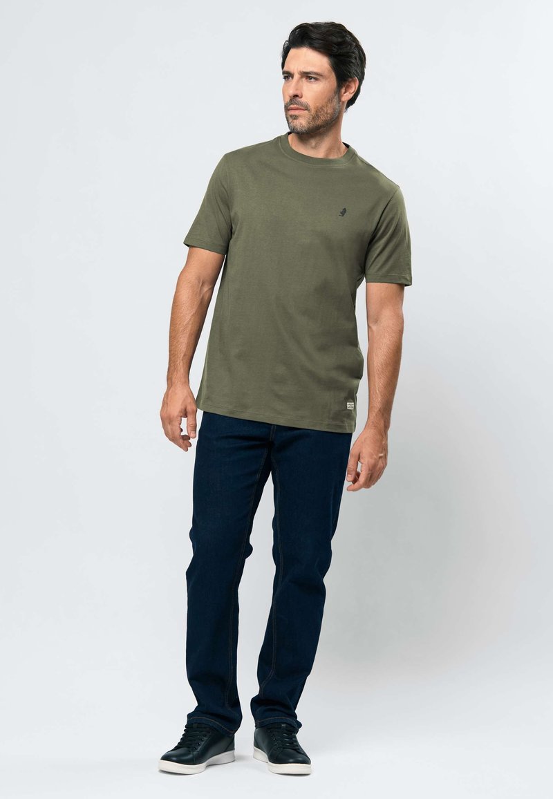 Homme debout portant un t-shirt à manches courtes vert olive, un jean bleu foncé et des baskets noires sur un fond clair uni.