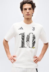 Νεαρός άνδρας που φοράει λευκό μπλουζάκι ποδοσφαίρου της Juventus με μαύρο αριθμό 10 και σχέδιο κλασικού αγάλματος, στέκεται με τα χέρια στη μέση.