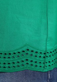 Zelený textilní top se zvlněným lemem a vystříhanými otvory. Hladká textura, lehký materiál a prodloužený šev na boku.