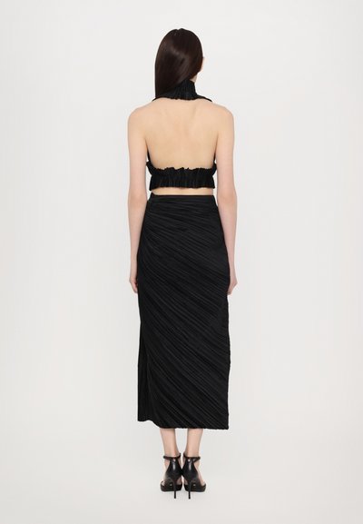 Black pleated two-piece outfit featuring a backless cropped top with ruffled edges and a mid-length skirt with diagonal pleats. High-heel shoes.
