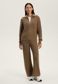 Trening din tricot texturat, de culoare maro, format dintr-o jachetă cu fermoar și pantaloni evazați. Prezintă un guler înalt și manșete ribbate. Purtați-l cu adidași albi.