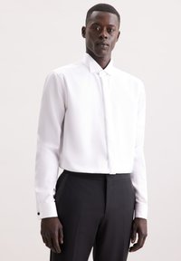 Camicia bianca con colletto alto, chiusura con un solo bottone e maniche lunghe, abbinata a pantaloni neri su misura con strisce laterali.