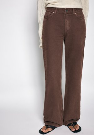 Pantalon droit marron porté avec des sandales noires et un haut beige à manches longues, visible de la taille aux pieds sur un fond uni.