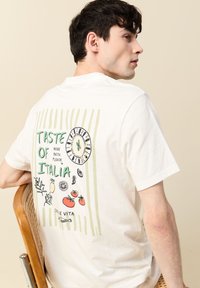 Valkoinen puuvillapaita, jossa on graafinen design selässä, jossa on vihreää ja mustaa tekstiä, ruokakuvastoja ja raitoja.