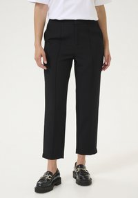 Pantaloni neri sartoriali a gamba dritta, con cuciture frontali e una superficie liscia. Abbinati a mocassini neri con accenti in metallo dorato.