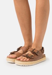Brown leather sandals with two adjustable buckles, thick woven jute midsole, and white rubber outsole, showing a textured finish.