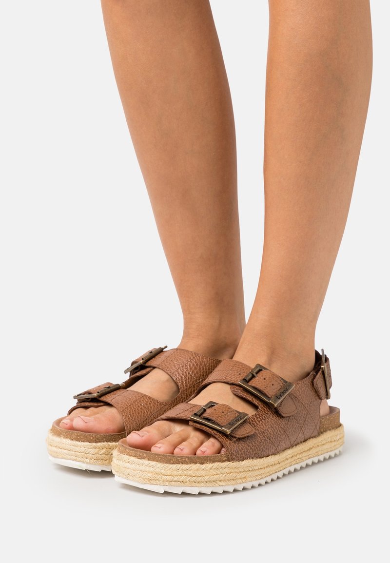 Brown leather sandals with two adjustable buckles, thick woven jute midsole, and white rubber outsole, showing a textured finish.