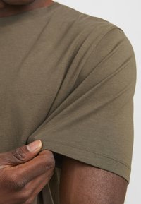 Olive green cotton t-shirt with a crew neckline, short sleeves, and visible stitching detail on the hem and seams. Hand is gripping the sleeve.