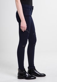 Tmavě modré slim džíny s roztrhanými kolenními panely a zipy, vyrobené z elastického denimového materiálu. V kombinaci s černými koženými kotníkovými botami.