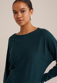 Turkizna pletena pulover s širokim izrezom in ohlapnim krojem, ki vključuje dolge rokave in mehak material. Srebrni ovalni uhani dopolnjujejo videz.