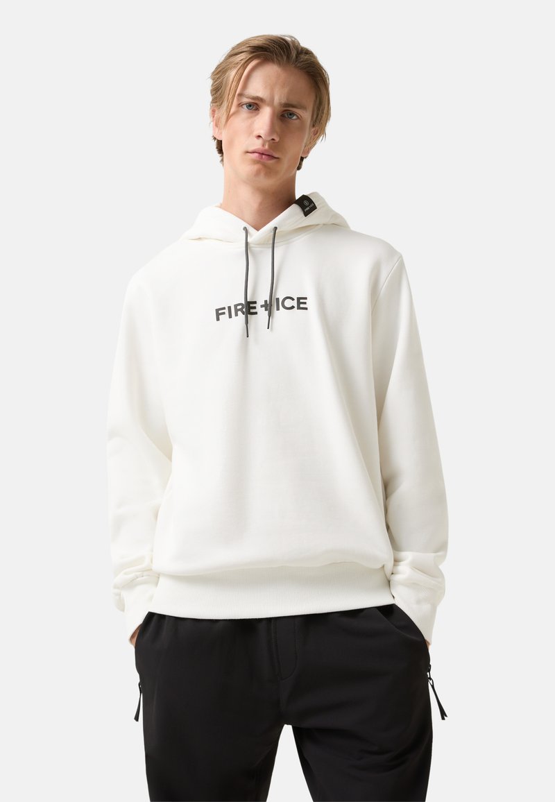 Młody mężczyzna z blond włosami, ubrany w biały hoodie z napisem "FIRE + ICE" i czarne spodnie, stoi z rękami w kieszeniach na jasnym tle.