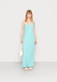 Vestido maxi sem mangas, azul claro, em um tecido suave e fluido, com decote drapeado e silhueta reta. Acompanhado de uma bolsa branca.