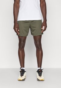 Αθλητικά σορτς ανδρικά σε ελαιόπρασινο, κατασκευασμένα από υφή ύφασμα. Συνδυασμένα με μαύρα και λευκά αθλητικά παπούτσια με πορτοκαλί λεπτομέρειες.