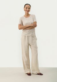 Haut à manches courtes à motifs floraux beige clair associé à un pantalon large beige clair et des chaussures foncées. Textures lisses tout au long.