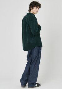Chemise en velours côtelé vert foncé avec col, manches longues et détail de patte de boutonnage à l'arrière, associée à un pantalon ample rayé bleu. Des chaussures plates viennent compléter la tenue.