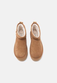 Botas de tobillo de ante marrón con un suave forro de piel de oveja blanca, punta redondeada y una lengüeta en la parte trasera. Exterior liso y acentos cosidos.
