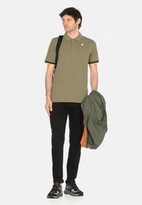 Polo verde oliva con maniche a righe gialle e nere, abbinato a jeans neri e una giacca verde. Il modello indossa sneakers nere.