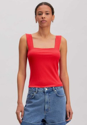 Jonge vrouw met gevlochten haar draagt een rood mouwloos topje met vierkante hals en een high-waist blauwe spijkerbroek tegen een effen achtergrond.