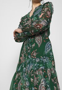 Mujer vestida con un vestido de manga larga de color verde con un patrón de paisley y detalles de volantes en los hombros, con los brazos cruzados, frente a un fondo sencillo.