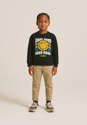 Zwarte sweatshirt met kleurrijke "Good Mood" tekst en een glimlachend gezicht graphic, gecombineerd met beige broek en grijze sneakers. Kind staat rechtop.