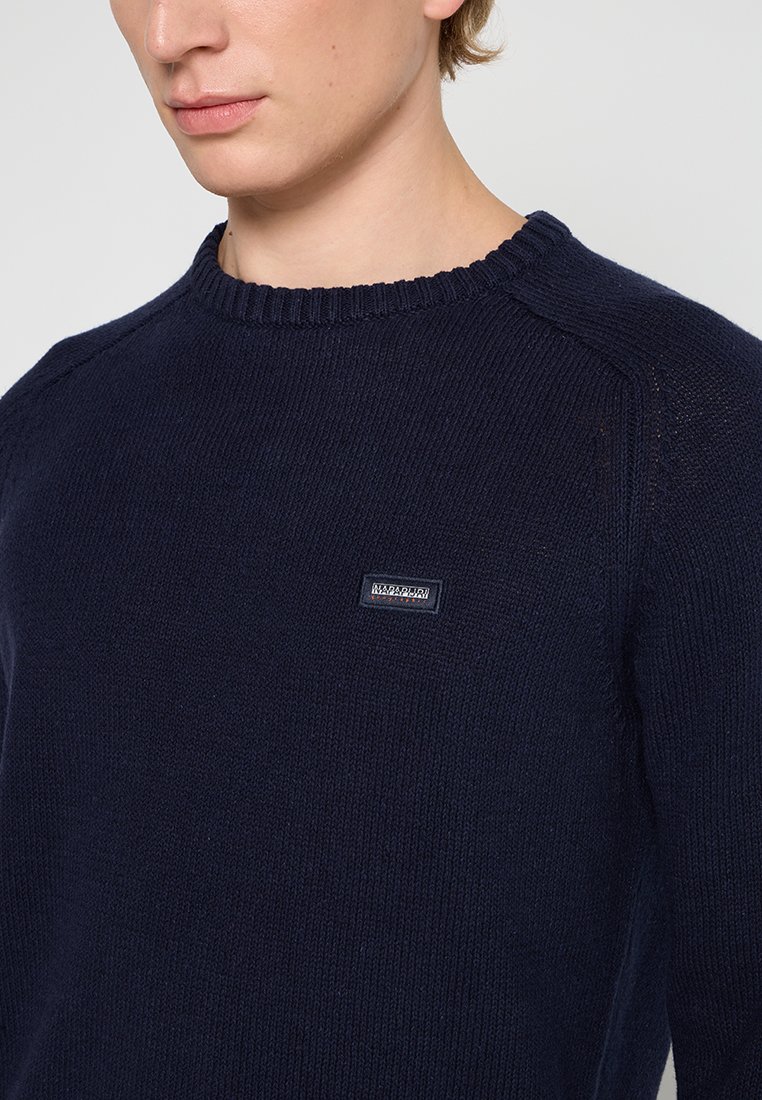 Pull en maille bleu marine avec un col rond côtelé, présentant un petit patch logo sur la poitrine, mettant en valeur une texture lisse et un design classique.