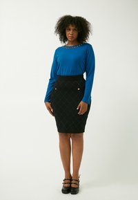 Langärmliges blaues Oberteil mit einem dekorativen Ausschnitt, kombiniert mit einem enganliegenden schwarzen Rock mit Diamantmustern und goldenen Knopfakzenten. Schwarze Absätze.