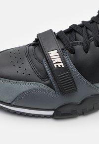 Sneakers Nike au caracteristici un superior din piele neagră și gri, o curea texturată cu logo și o talpă din cauciuc cu accente albe și detalii perforate.