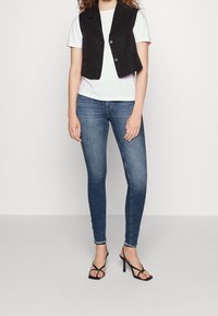 Chaleco negro recortado con dos botones sobre una camiseta blanca, combinado con jeans skinny azules y sandalias negras con tiras.