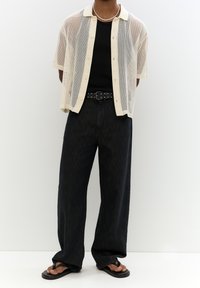 Krótki beżowy koszulka z siateczki na czarnym dopasowanym topie, w zestawieniu z luźnymi czarnymi dżinsami i czarnymi klapkami. Podkreślona ćwiekowanym paskiem.