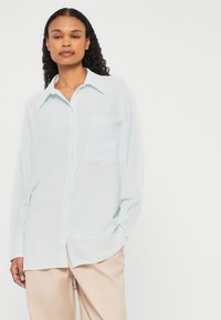 Hellblaues, langärmliges Hemd mit durchgeknöpfter Vorderseite, einer aufgesetzten Tasche und bequemer Passform. Der Stoff hat ein subtile Struktur.