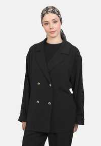 Zwarte double-breasted blazer met gouden knopen, relaxte pasvorm, brede revers en lange mouwen; gedragen over een zwart top.
