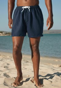 Tengerészkék fürdőnadrág elasztikus derékkal, fehér húzózsinórral és oldalsó varrással, áll egy strandnál, a háttérben sekély víz látható.