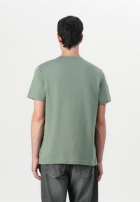 Tricou din bumbac verde, cu guler rotund și mâneci scurte, având un tiv drept și o textură netedă, mată. Design simplu, fără grafice.