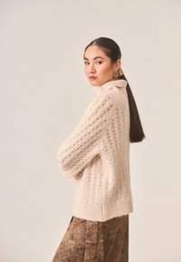 Suéter de punto color crema con cuello alto acanalado, que presenta un patrón de tejido abierto. Se combina con una falda marrón con estampado.