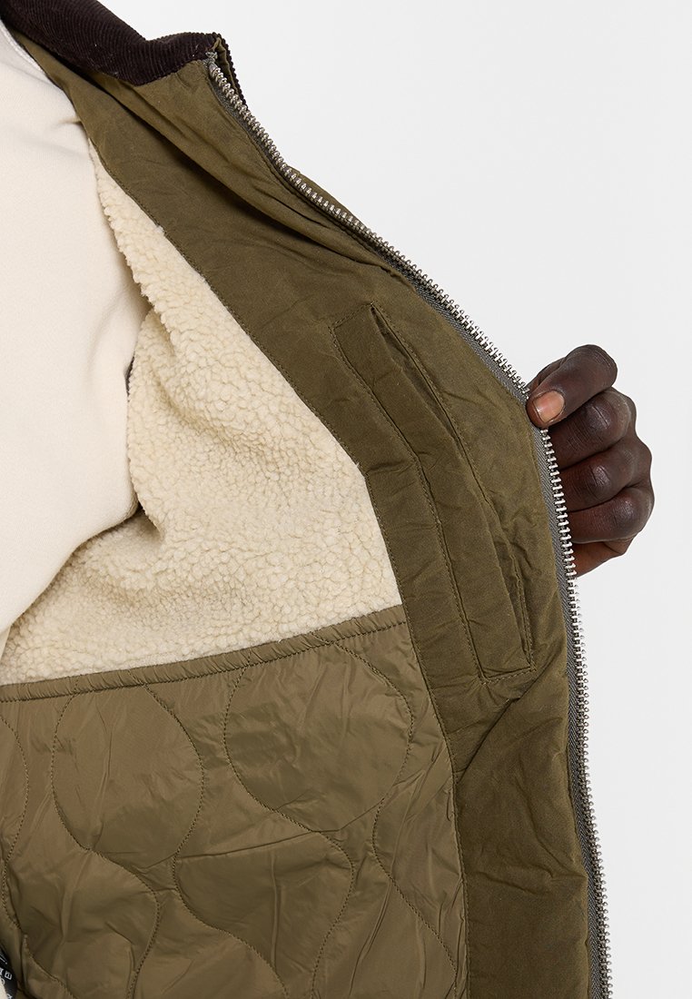 Ogrodowy zielony pikowany płaszcz wewnątrz z kremowym polarem, posiadający płaską kieszeń i srebrny zamek. Gładka struktura na całej powierzchni.