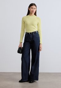 Κίτρινη ριγέ μπλούζα με μακριά μανίκια, συνδυασμένη με σκούρο μπλε φαρδιά τζιν παντελόνια. Το μοντέλο κρατά μια μικρή μαύρη τσάντα, στέκεται σε ουδέτερο επιφάνεια με υφή.